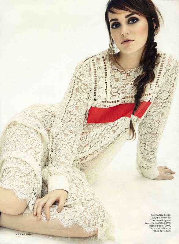 Woman poses in a long-sleeved lace dress featuring a bold red stripe across the chest, seated on a light neutral surface indoors.