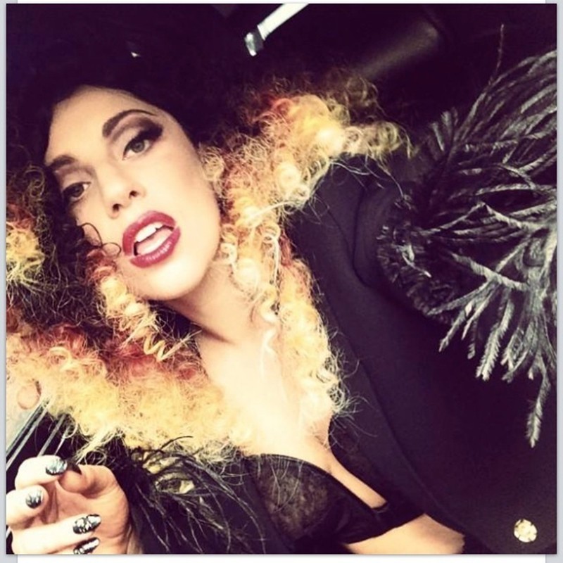 Close-up selfie of a stylish woman with curly blonde and dark hair, bold makeup, and an elegant black jacket with feather details.