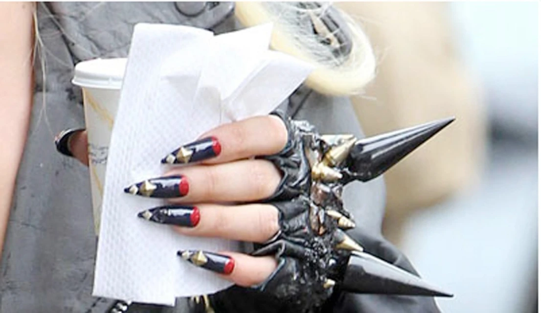Close-up of a woman's hand wearing a black spiked glove and long nails, holding a beverage cup and tissues, against a blurred background.
