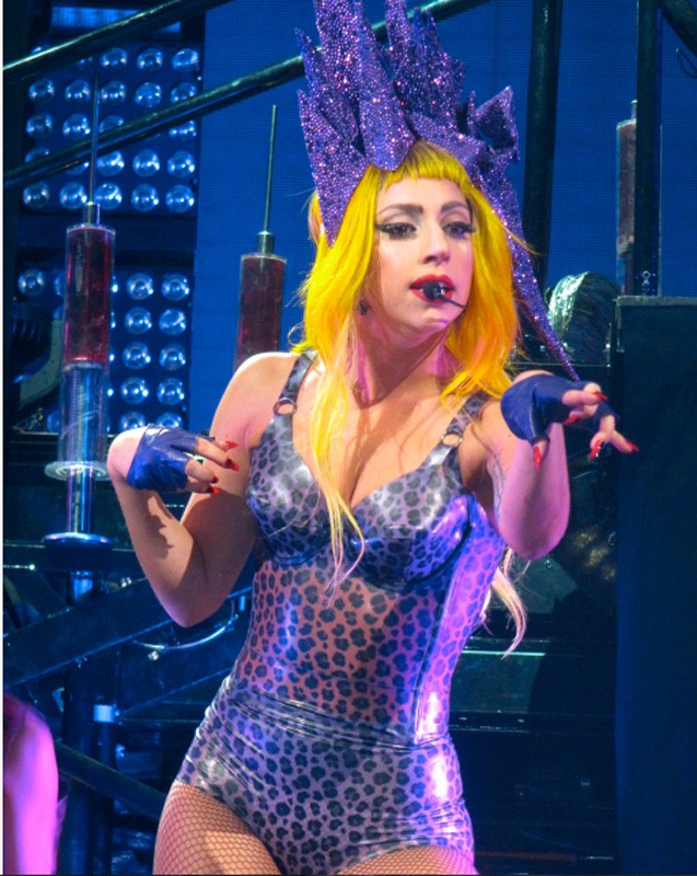 Lady Gaga performs on stage with a shiny, leopard print bodysuit and a purple headdress, set against a blue and metallic background.
