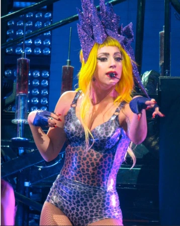 Lady Gaga performs on stage with a shiny, leopard print bodysuit and a purple headdress, set against a blue and metallic background.