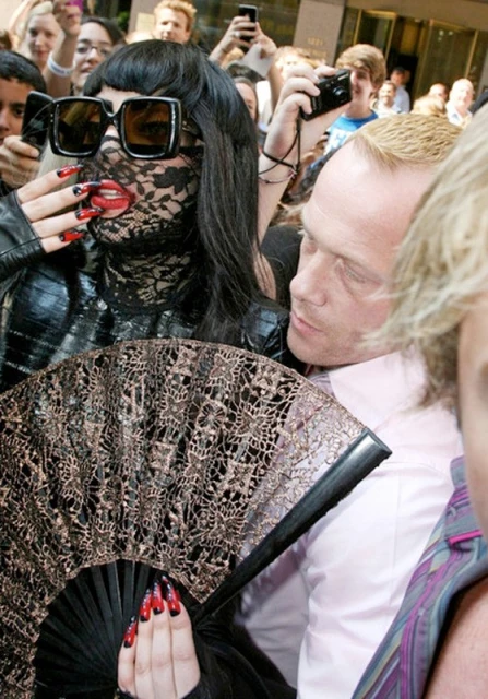 Singer Lady Gaga wearing a black lace face covering, large sunglasses, and holding a fan, with crowds and a building in the background.