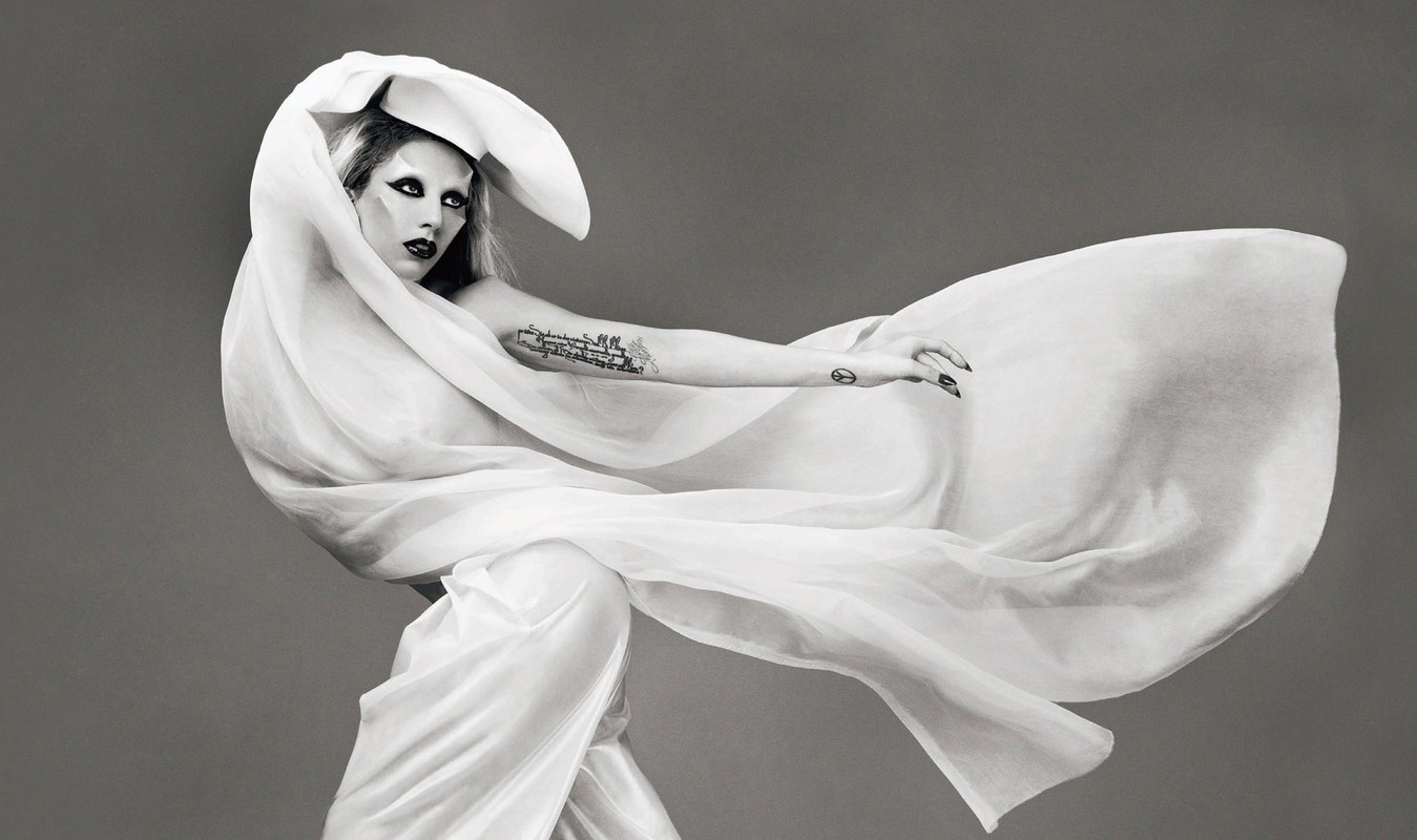 Lady Gaga wearing a draped white fabric wrap and sculptural headpiece against a grey background, styled with dark eye makeup and nail art.
