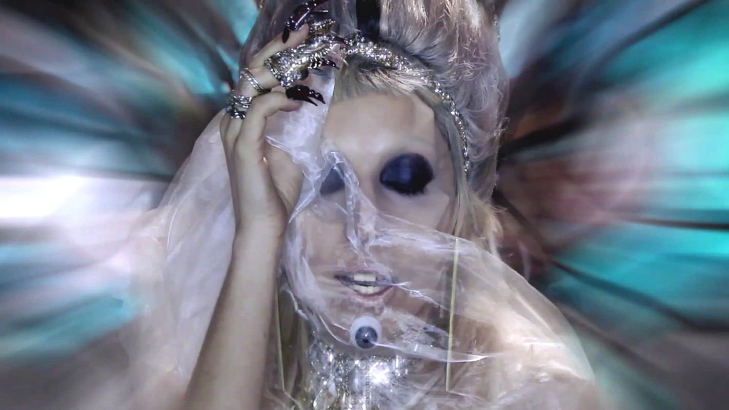 Close-up of a performer with a plastic veil and dramatic makeup, accessorized with jewelry and a headpiece against a blurred teal background.