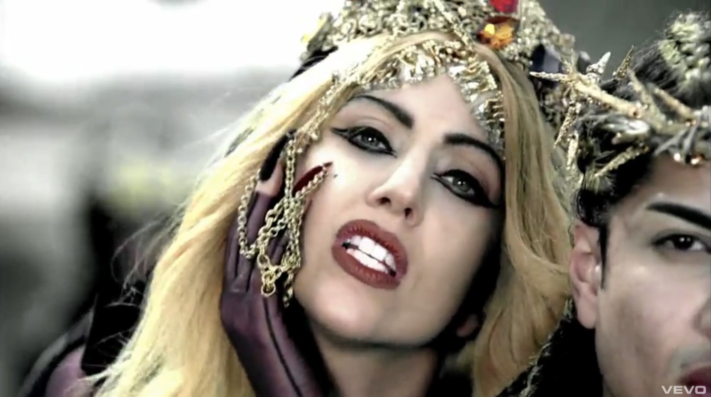 Close-up of woman with blond hair, dark eye makeup, and a jeweled headpiece, next to a person with a similar crown, on a grey background.