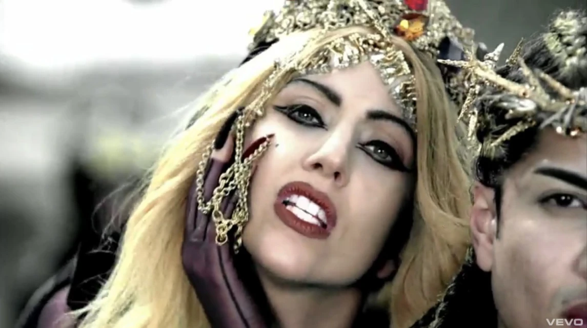 Close-up of woman with blond hair, dark eye makeup, and a jeweled headpiece, next to a person with a similar crown, on a grey background.