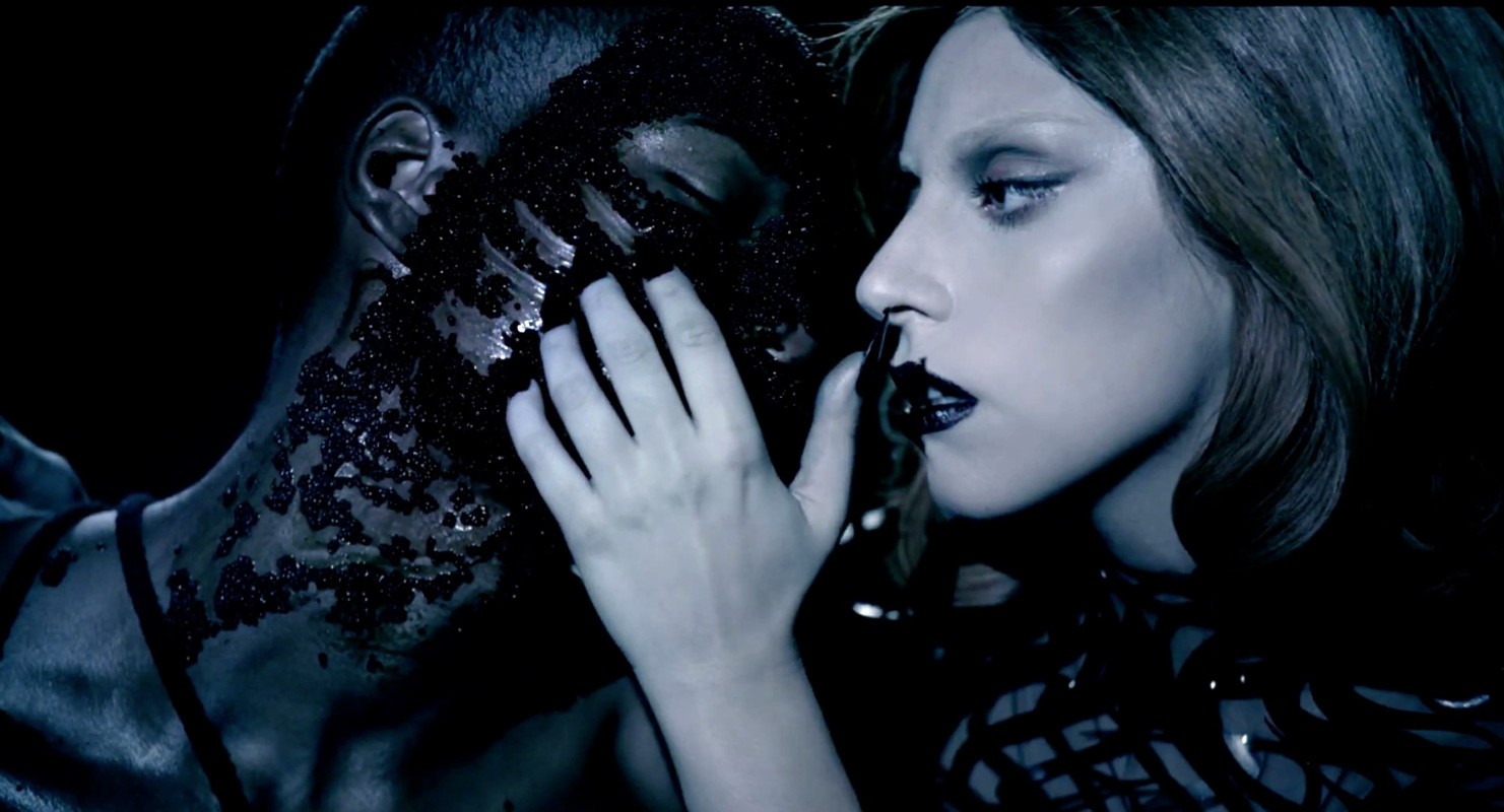 Close-up of pop singer Lady Gaga touching the face of a dark-skinned man covered with small black objects in a dark environment.