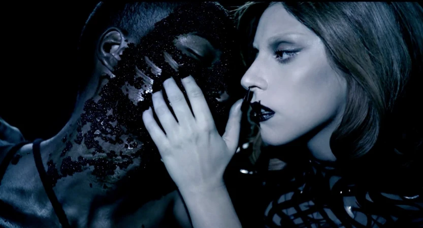Close-up of pop singer Lady Gaga touching the face of a dark-skinned man covered with small black objects in a dark environment.