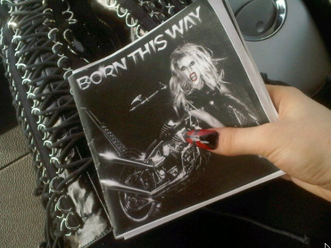 Close-up of a hand holding a 'Born This Way' album cover with an image of Lady Gaga on a motorcycle, with a black lace-up garment nearby.
