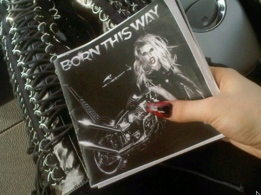 Close-up of a hand holding a 'Born This Way' album cover with an image of Lady Gaga on a motorcycle, with a black lace-up garment nearby.