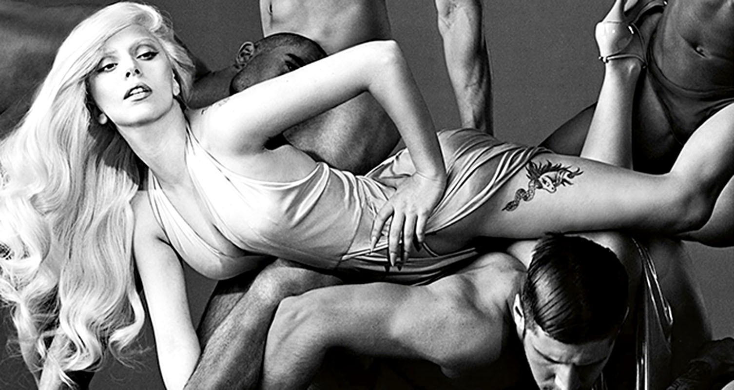 Monochrome image of Lady Gaga reclining amidst shirtless men, showcasing a tattoo, high heels, and an elegant dress in a striking studio shot.