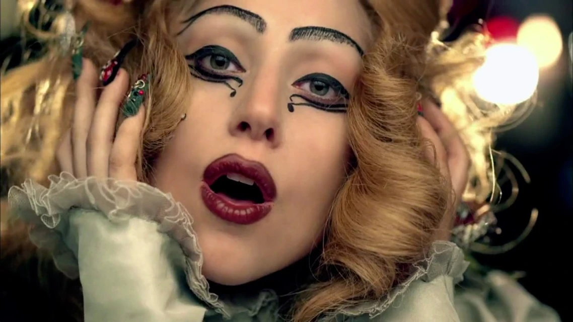 Dramatic close-up of a blonde-haired woman with bold makeup, including exaggerated eyeliner and red lipstick, set against a dark background.