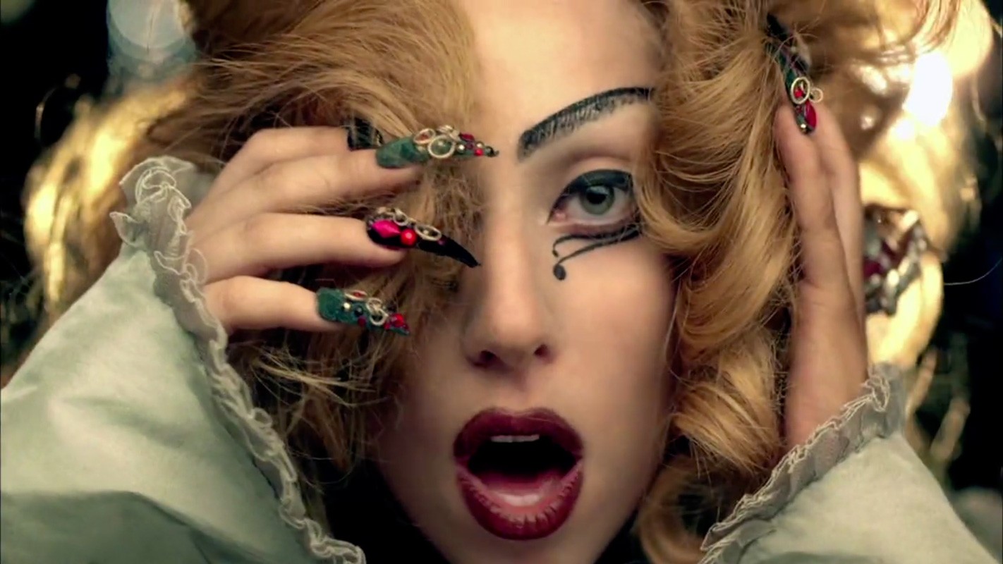 Close-up portrait of a woman with dramatic eye makeup, holding her hands to her blonde hair, featuring ornate jewelled long nails.