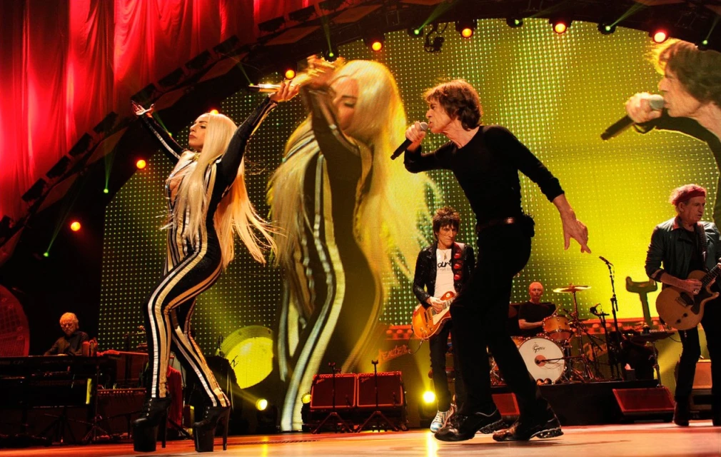Live concert performance featuring Lady Gaga and The Rolling Stones on stage. Two adult female performers and a group of adult male musicians.
