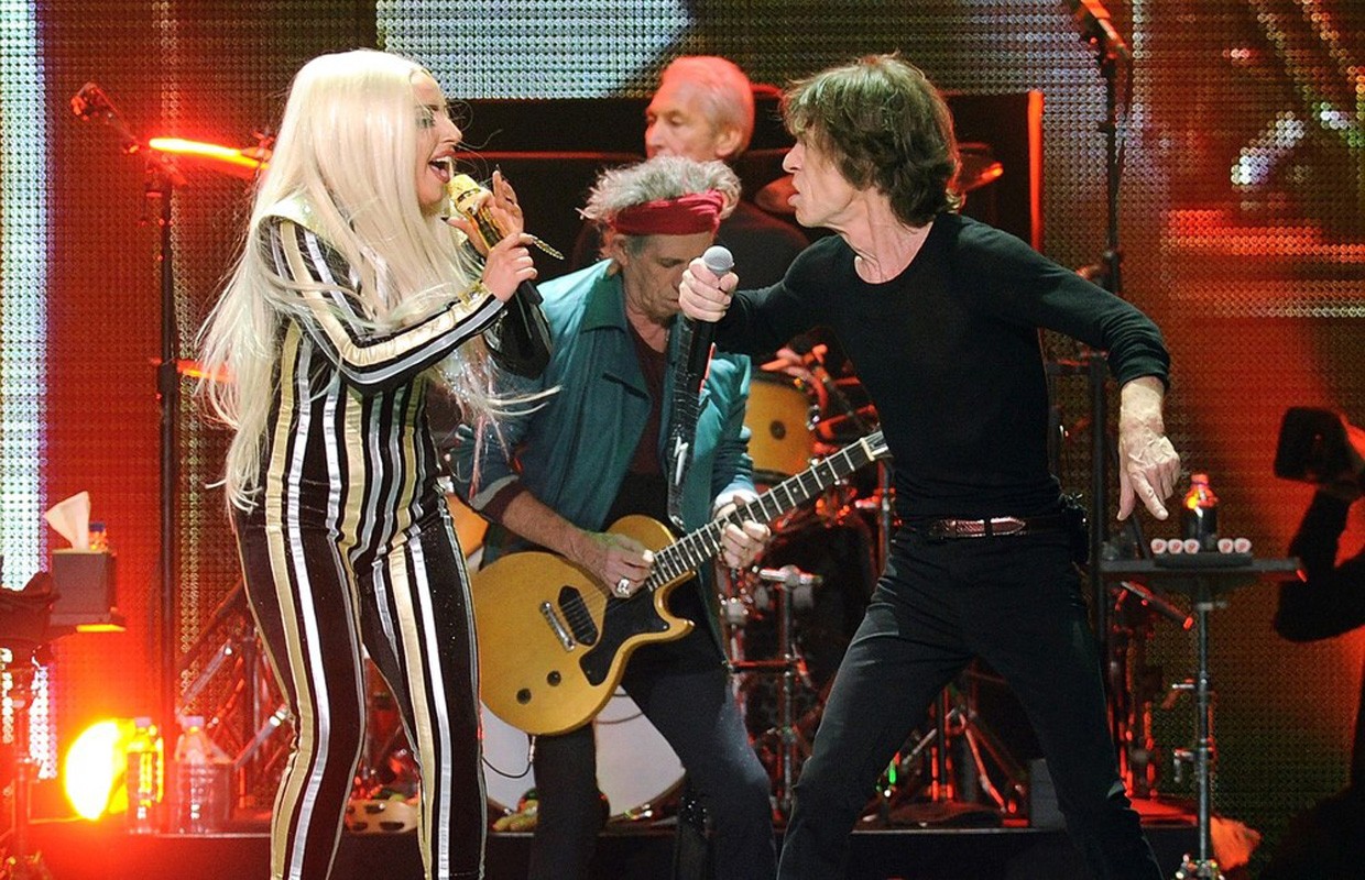 Performance shot of Lady Gaga and Mick Jagger singing on stage, accompanied by Keith Richards on guitar during a rock concert.