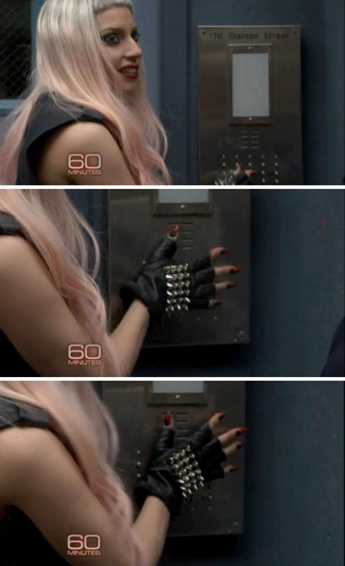 Close-up shot of a woman with pink hair wearing a studded black glove reaching for an elevator button indoors with gray walls.