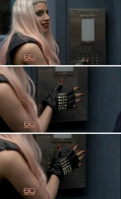 Close-up shot of a woman with pink hair wearing a studded black glove reaching for an elevator button indoors with gray walls.
