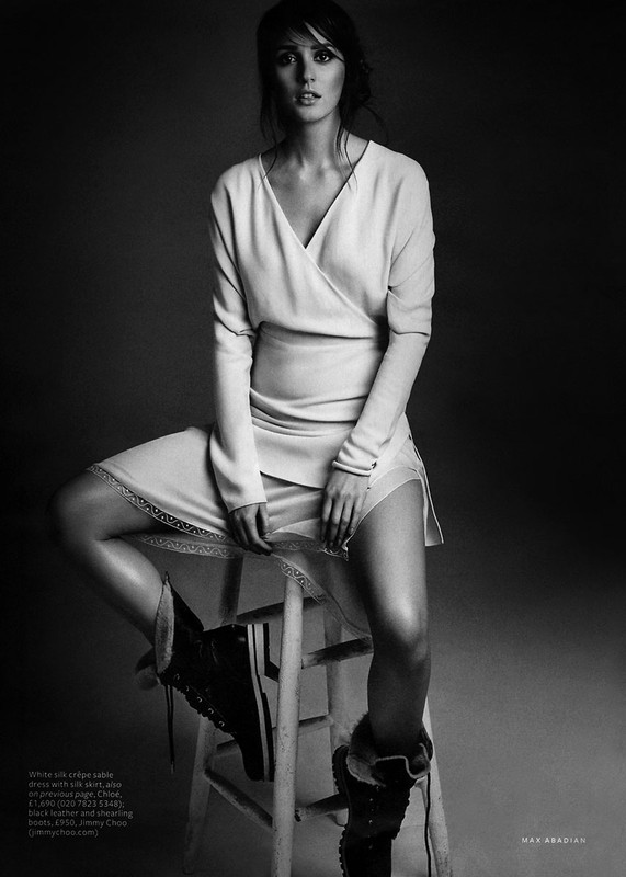 Black and white shot of a woman posing in a dress and boots, sitting on a stool. Studio setting with soft lighting.