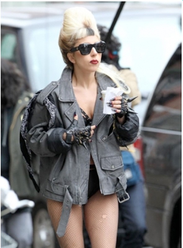 Blonde woman with beehive hairstyle and sunglasses wears a gray leather jacket with spiked gloves and fishnet stockings in urban setting.