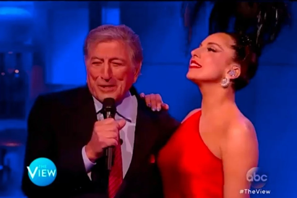 Tony Bennett and Lady Gaga perform on The View, captured in a medium shot with a blue background. The pair sings into a microphone.