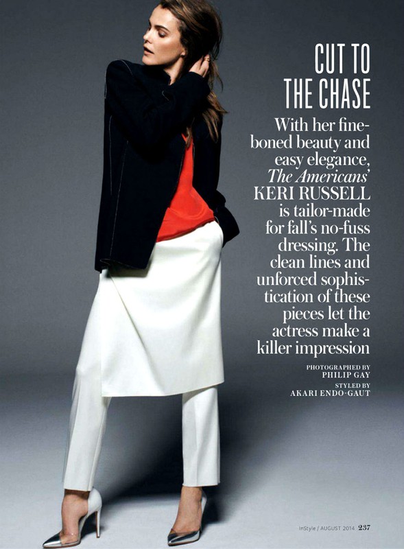 Fashion editorial shot of a woman in a black blazer, orange top, and white pants, posing against a grey backdrop in a studio setting.