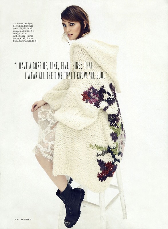Brunette woman poses on a white stool in a studio wearing a hooded, cable-knit cardigan and black boots in a fashion editorial shot.