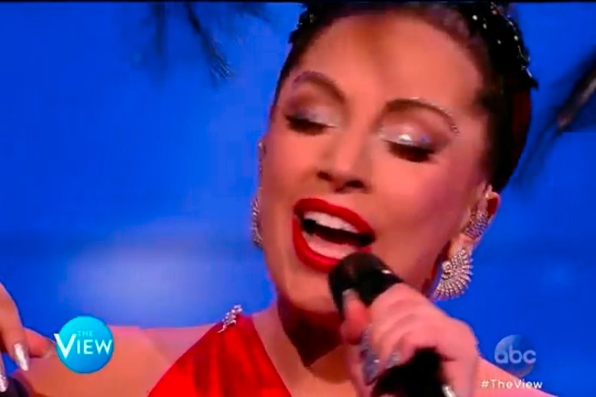 Singer on stage with bright red lipstick and ornate earrings, holding a microphone in front of a blue background.