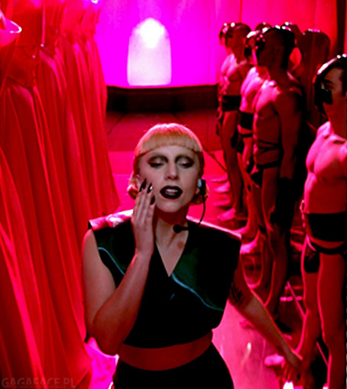 Lady Gaga performs on stage with dancers in matching outfits, bathed in pink and red light. She wears a black outfit with a microphone headset.