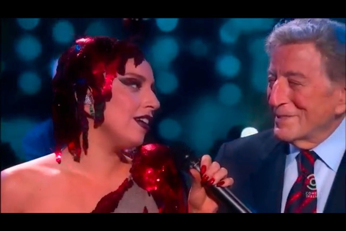 Lady Gaga and Tony Bennett perform on stage; Gaga wears a red sequin outfit, while Bennett is in a suit and tie; blue bokeh background.