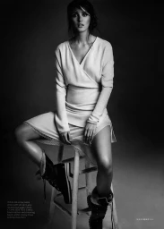 Woman seated on a wooden stool wearing a light-coloured wrap top, skirt, and dark boots against a dark background