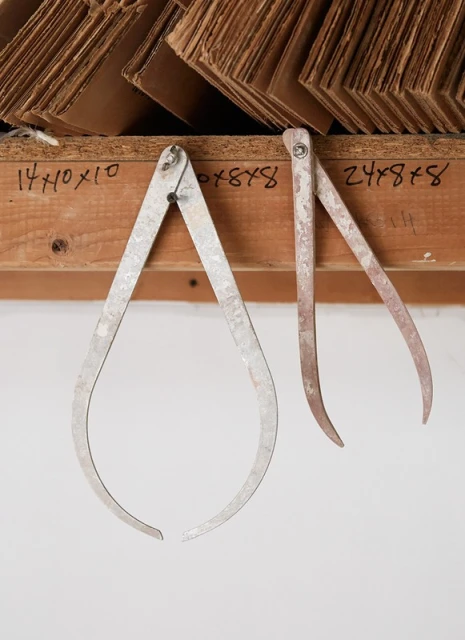 Two metal calipers hang from a wooden shelf with cardboard sheets stacked on top, set against a plain white wall indoors.