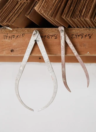 Two metal calipers hang from a wooden shelf with cardboard sheets stacked on top, set against a plain white wall indoors.