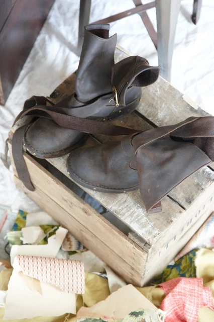 Rustic brown leather ankle boots with straps on a weathered wooden crate with textile scraps, showcasing handcrafted footwear.