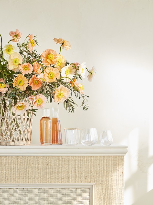 Elegant arrangement of peach and white flowers in a woven vase, accompanied by clear glass carafes and glasses on a white shelf.