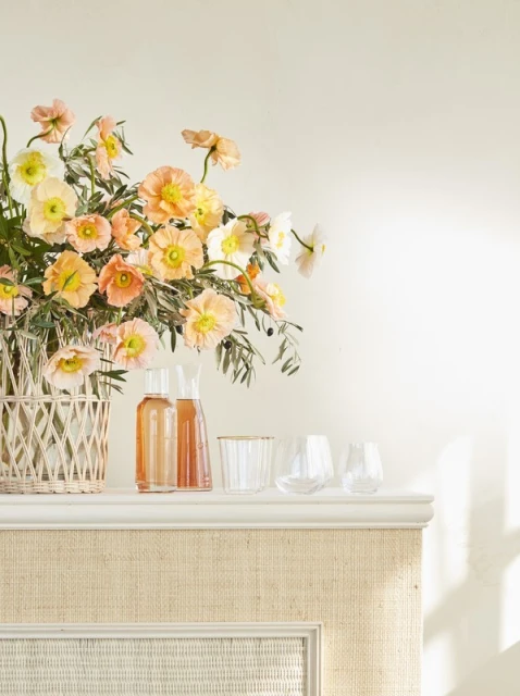 Elegant arrangement of peach and white flowers in a woven vase, accompanied by clear glass carafes and glasses on a white shelf.