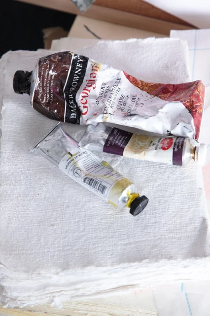 Two tubes of artist's paint laying on top of a stack of textured white paper in a close-up studio shot.