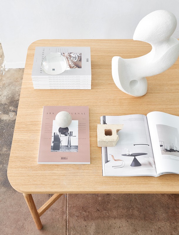 Wooden table surface featuring art journals, a stack of magazines, a white sculpture, and various decorative objects in an interior setting.