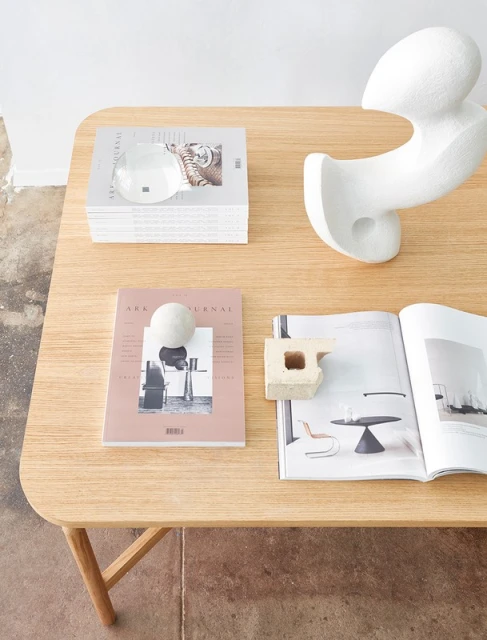Wooden table surface featuring art journals, a stack of magazines, a white sculpture, and various decorative objects in an interior setting.