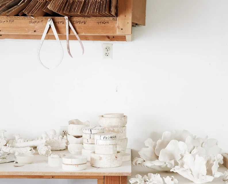 Still life of handmade ceramic bowls and art tools on a wooden table in a white-walled studio, emphasizing craftsmanship and art creation.