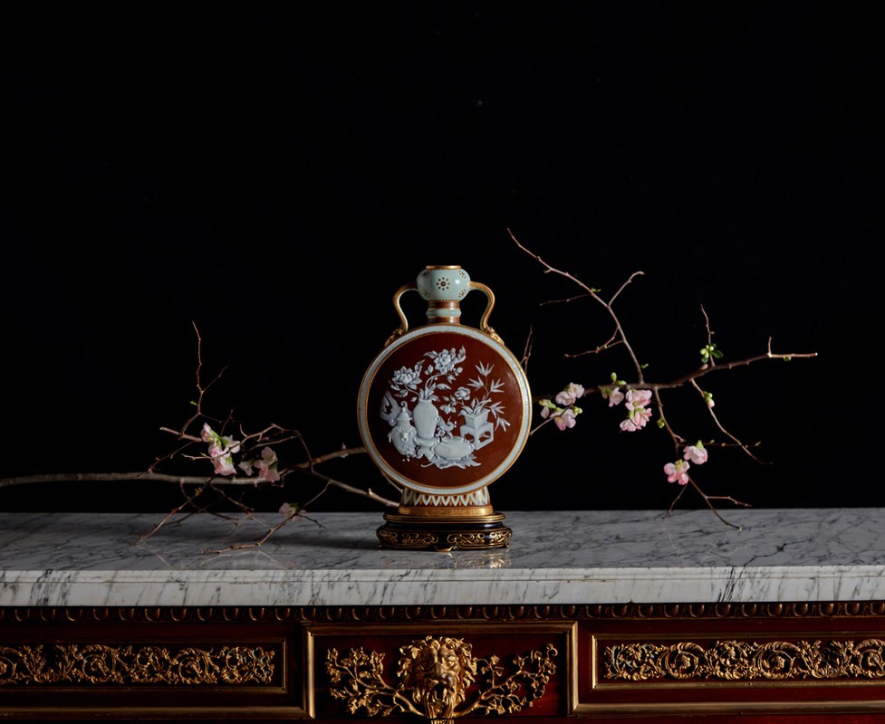 Still life of decorative vase with white floral design, branches with pink blossoms on a marble surface against black backdrop.