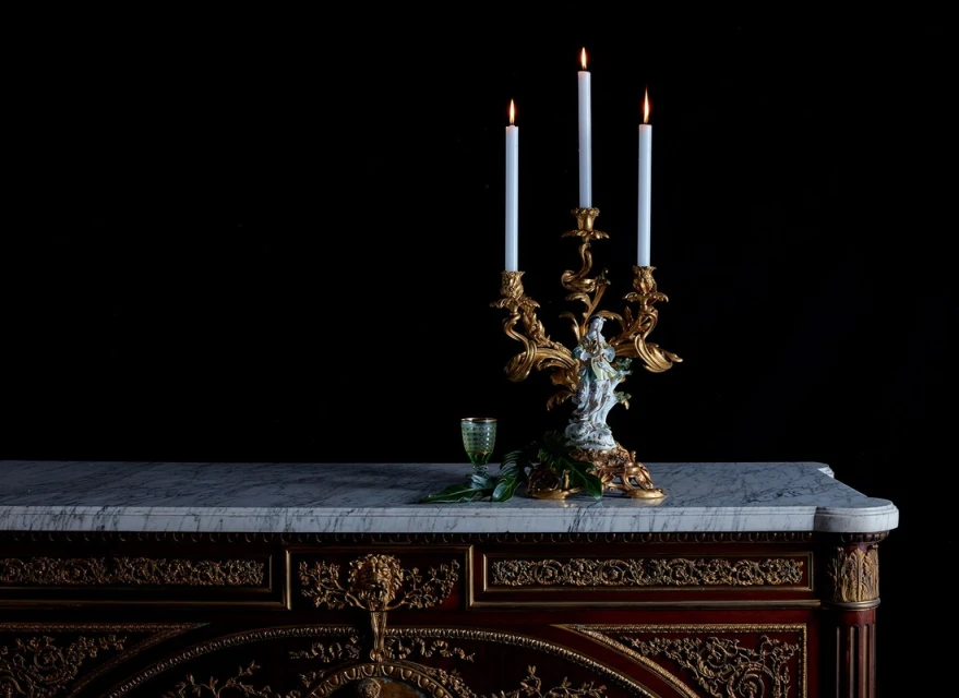 Ornate candelabra with lit candles displayed on a marble-topped credenza against a dark background, casting a warm glow.