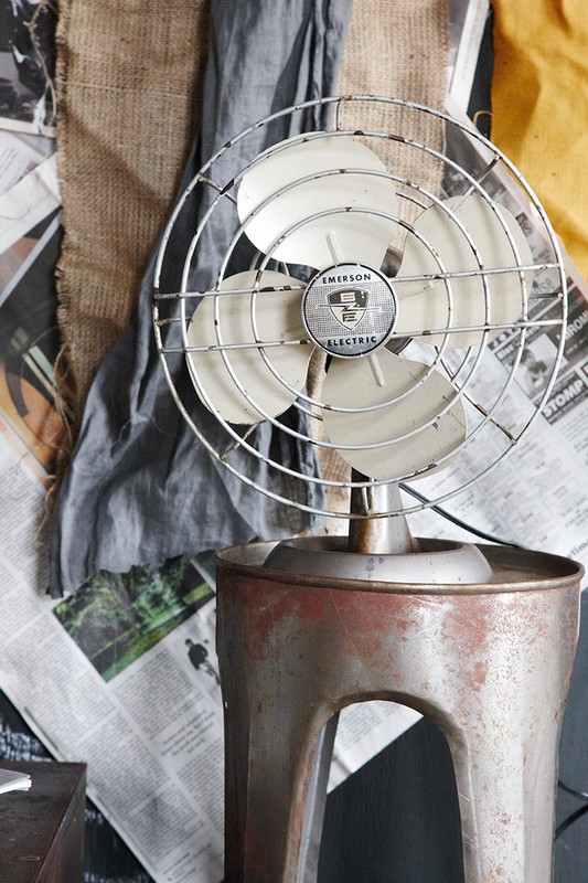 Vintage Emerson Electric fan on a metal stand is displayed in front of newspaper and fabric scraps in a studio setting.