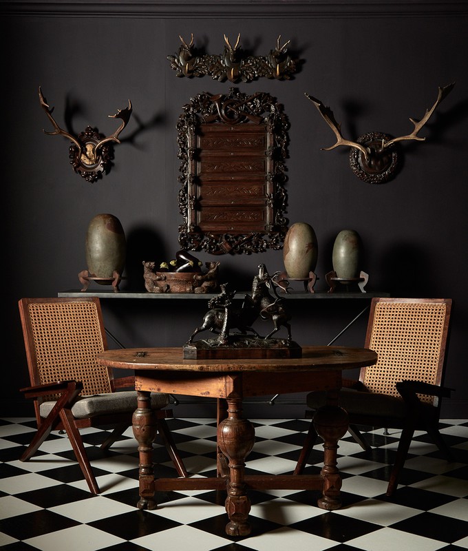 Interior home view features a round wooden table, sculpture, chairs, vases, & wall decor of mounted antlers on black and white checkered floor.