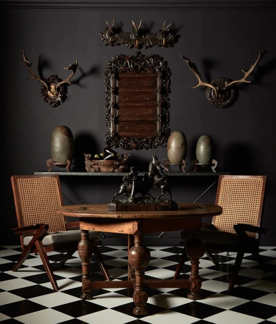 Interior home view features a round wooden table, sculpture, chairs, vases, & wall decor of mounted antlers on black and white checkered floor.