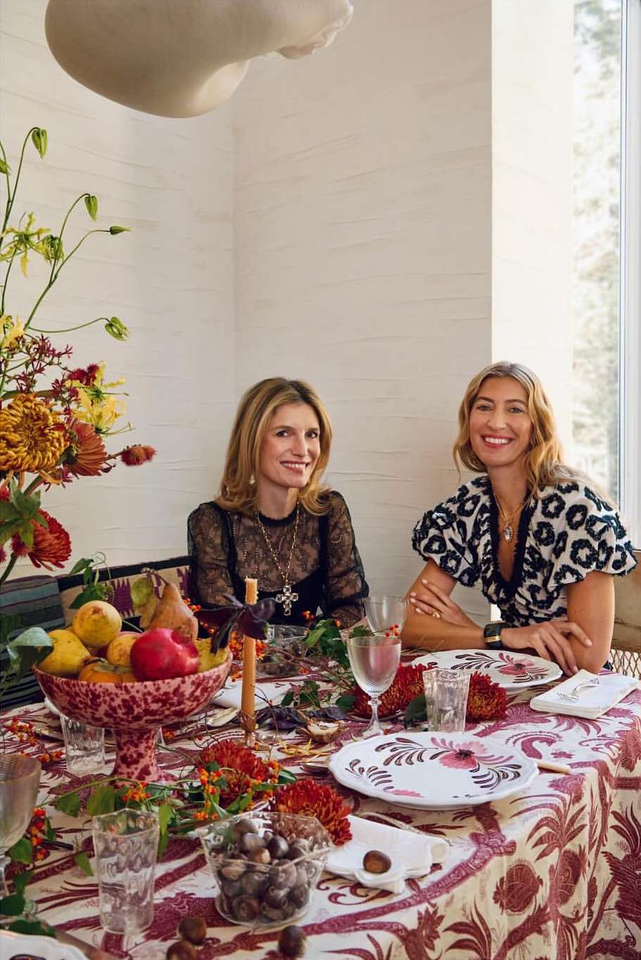 Two adult women sit at a dining table with patterned tablecloth and floral arrangement centerpiece, with floral tableware and decor.