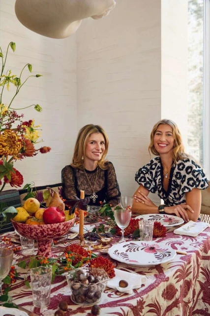 Two adult women sit at a dining table with patterned tablecloth and floral arrangement centerpiece, with floral tableware and decor.