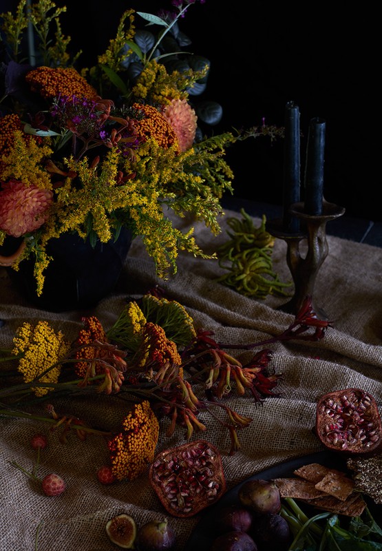 Dramatic floral arrangement with dark candles, pomegranates, and figs on burlap in moody lighting against a dark background.