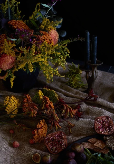 Dramatic floral arrangement with dark candles, pomegranates, and figs on burlap in moody lighting against a dark background.
