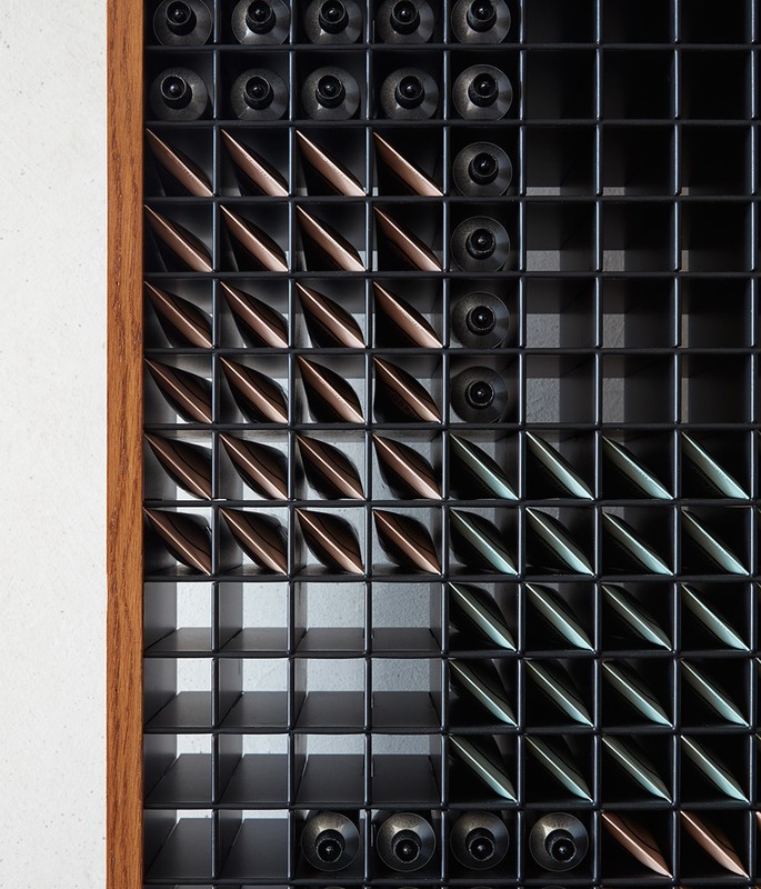 Geometric pattern art installation in a grid with brown, bronze, and black elements, framed by a light wood trim, on a white wall.