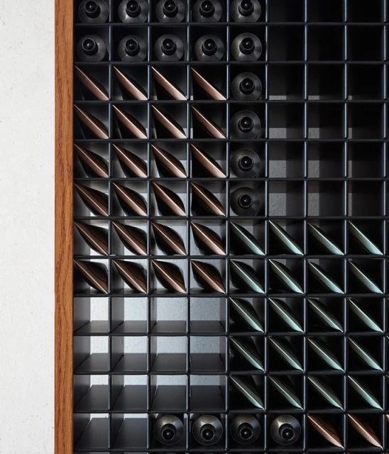 Geometric pattern art installation in a grid with brown, bronze, and black elements, framed by a light wood trim, on a white wall.